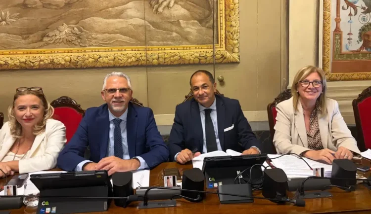 Quattro persone sedute a un banco con microfoni e documenti, in un’aula dove si discute di aiuti alle imprese siciliane
