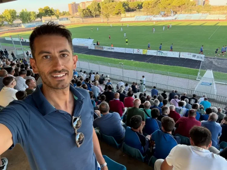 Simone Digrandi in un selfie in tribuna durante una partita di calcio, con il pubblico sugli spalti alle sue spalle