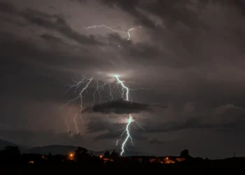 Fulmini da un temporale notturno sopra un paese illuminato, con colline sullo sfondo, simbolo dell’allerta gialla a Ragusa
