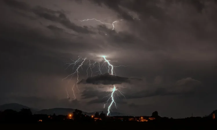 Fulmini da un temporale notturno sopra un paese illuminato, con colline sullo sfondo, simbolo dell’allerta gialla a Ragusa