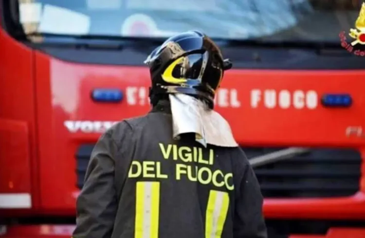 Vigile del fuoco di spalle con casco e giacca con la scritta «Vigili del Fuoco» davanti a un camion rosso dei pompieri