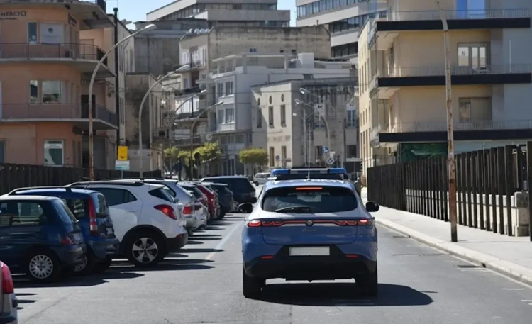Auto della Polizia di Stato in servizio in una strada di Ragusa, tra auto parcheggiate e palazzi del centro