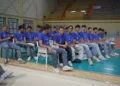 Giocatori della Volley Modica in maglia blu seduti in fila a bordo campo in un palazzetto con le tribune vuote