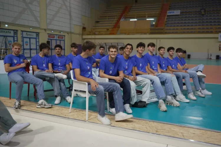 Giocatori della Volley Modica in maglia blu seduti in fila a bordo campo in un palazzetto con le tribune vuote