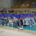Giocatori della Volley Modica in maglia blu seduti in fila a bordo campo in un palazzetto con le tribune vuote