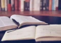 Libro aperto con una penna appoggiata su un quaderno di appunti scritti a mano, con una fila di libri sullo sfondo