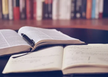 Libro aperto con una penna appoggiata su un quaderno di appunti scritti a mano, con una fila di libri sullo sfondo