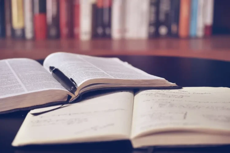 Libro aperto con una penna appoggiata su un quaderno di appunti scritti a mano, con una fila di libri sullo sfondo
