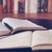 Libro aperto con una penna appoggiata su un quaderno di appunti scritti a mano, con una fila di libri sullo sfondo