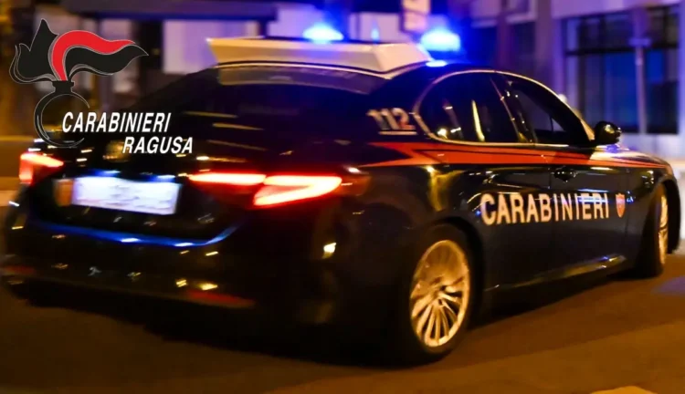 Auto dei Carabinieri vista da dietro, di notte in una strada, con lampeggianti blu accesi e logo Carabinieri Ragusa in sovrimpressione