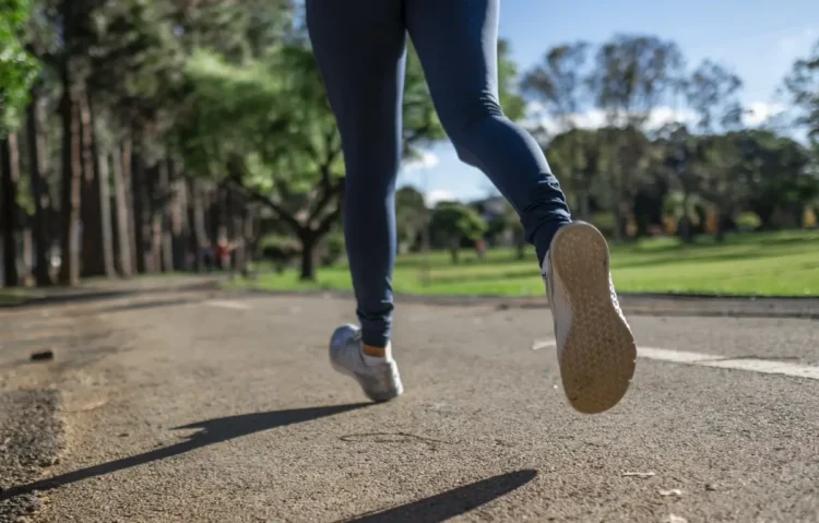 Gambe di una persona in leggings blu e scarpe da ginnastica che corre su un viale asfaltato in un parco alberato