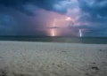 Fulmini cadono sul mare all’orizzonte sotto nuvole scure, con una spiaggia di sabbia chiara in primo piano durante l’allerta meteo