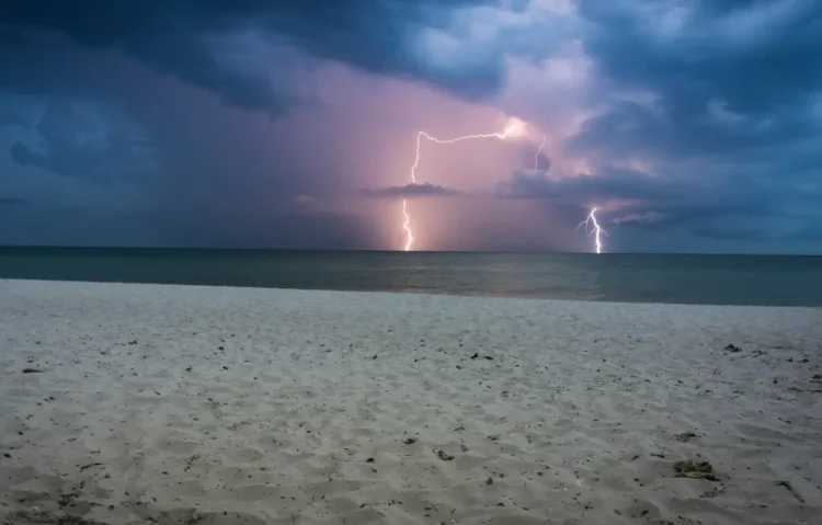 Fulmini cadono sul mare all’orizzonte sotto nuvole scure, con una spiaggia di sabbia chiara in primo piano durante l’allerta meteo