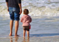Un adulto e una bambina in felpa rosa, di spalle, in riva al mare: foto simbolica sull’assegno unico per i figli