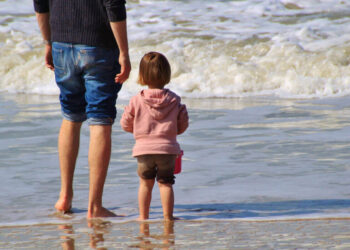 Un adulto e una bambina in felpa rosa, di spalle, in riva al mare: foto simbolica sull’assegno unico per i figli