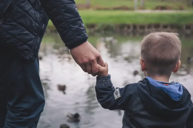 Un adulto tiene per mano un bambino ripreso di spalle sulla riva di uno stagno con le anatre, foto per l'assegno unico