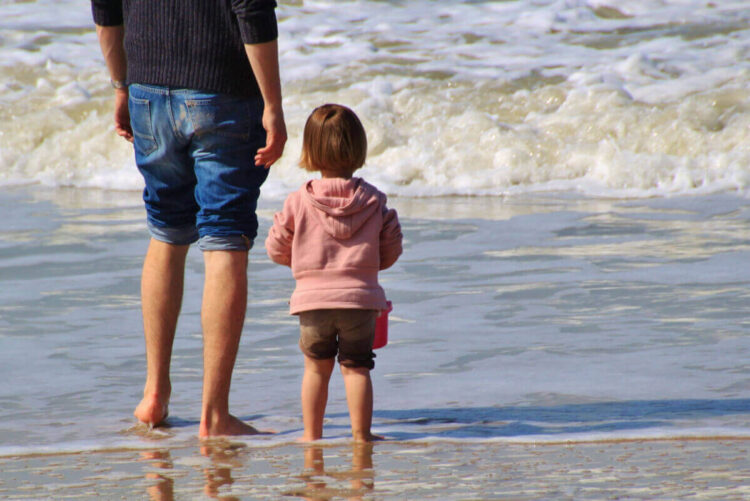 Un adulto e una bambina in felpa rosa, di spalle, in riva al mare: foto simbolica sull’assegno unico per i figli