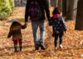 Un genitore cammina di spalle in un viale alberato coperto di foglie autunnali, tenendo per mano due bambini piccoli