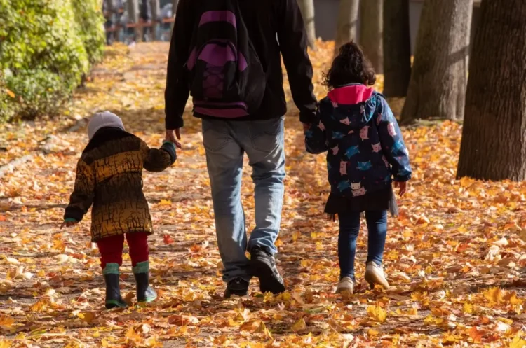 Un genitore cammina di spalle in un viale alberato coperto di foglie autunnali, tenendo per mano due bambini piccoli