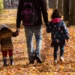 Un genitore cammina di spalle in un viale alberato coperto di foglie autunnali, tenendo per mano due bambini piccoli