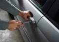Mani di un uomo in maglia grigia che forzano con un cacciavite la serratura della portiera di un’auto argentata