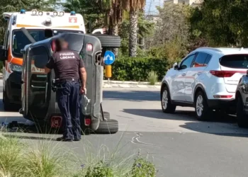 Auto ribaltata su un fianco in una strada di Comiso, un agente della Polizia locale accanto e un'ambulanza del 118 dietro