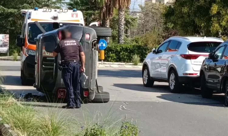 Auto ribaltata su un fianco in una strada di Comiso, un agente della Polizia locale accanto e un'ambulanza del 118 dietro