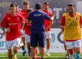 Giocatori del Modica Calcio in maglia rossa che corrono in riscaldamento su un campo sintetico, con un membro dello staff di spalle