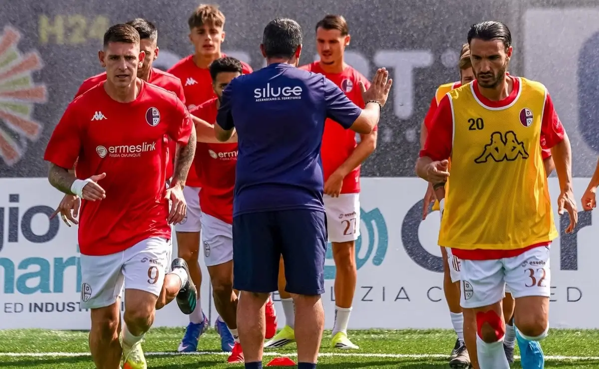 Giocatori del Modica Calcio in maglia rossa che corrono in riscaldamento su un campo sintetico, con un membro dello staff di spalle
