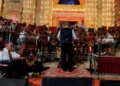 La banda di Hamrun in divisa bianca e rossa suona su un palco davanti alla facciata illuminata di una chiesa, con il direttore sul podio