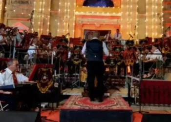 La banda di Hamrun in divisa bianca e rossa suona su un palco davanti alla facciata illuminata di una chiesa, con il direttore sul podio