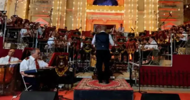 La banda di Hamrun in divisa bianca e rossa suona su un palco davanti alla facciata illuminata di una chiesa, con il direttore sul podio