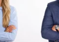 Una donna in camicia azzurra e un uomo in giacca blu, separati e a braccia conserte, ai due lati opposti dell’immagine