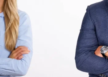 Una donna in camicia azzurra e un uomo in giacca blu, separati e a braccia conserte, ai due lati opposti dell’immagine