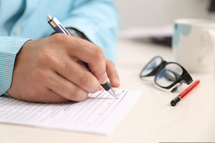 Mano che compila un modulo con la penna su una scrivania, accanto a occhiali e tazza: pratiche per bonus assunzioni