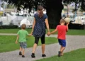Una mamma di spalle cammina per mano con due bambini su un vialetto di ghiaia in un parco, vicino a un porto con barche a vela