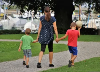 Una mamma di spalle cammina per mano con due bambini su un vialetto di ghiaia in un parco, vicino a un porto con barche a vela