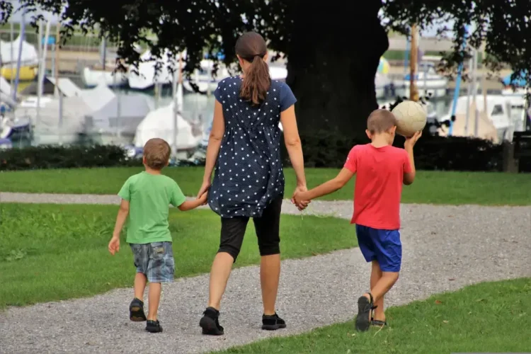 Una mamma di spalle cammina per mano con due bambini su un vialetto di ghiaia in un parco, vicino a un porto con barche a vela