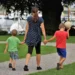 Una mamma di spalle cammina per mano con due bambini su un vialetto di ghiaia in un parco, vicino a un porto con barche a vela