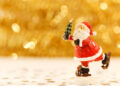 Statuina in legno dipinto di Babbo Natale sui pattini con un alberello in mano, su fondo dorato sfocato e coriandoli a stella