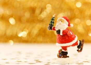 Statuina in legno dipinto di Babbo Natale sui pattini con un alberello in mano, su fondo dorato sfocato e coriandoli a stella