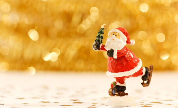 Statuina in legno dipinto di Babbo Natale sui pattini con un alberello in mano, su fondo dorato sfocato e coriandoli a stella
