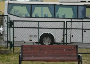 Un pullman per il trasporto passeggeri, con i sedili vuoti, fermo dietro una recinzione metallica verde; davanti, una panchina di legno