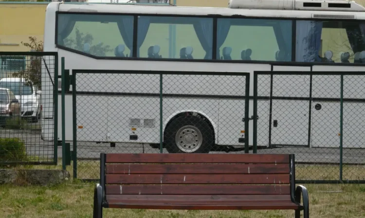 Un pullman per il trasporto passeggeri, con i sedili vuoti, fermo dietro una recinzione metallica verde; davanti, una panchina di legno