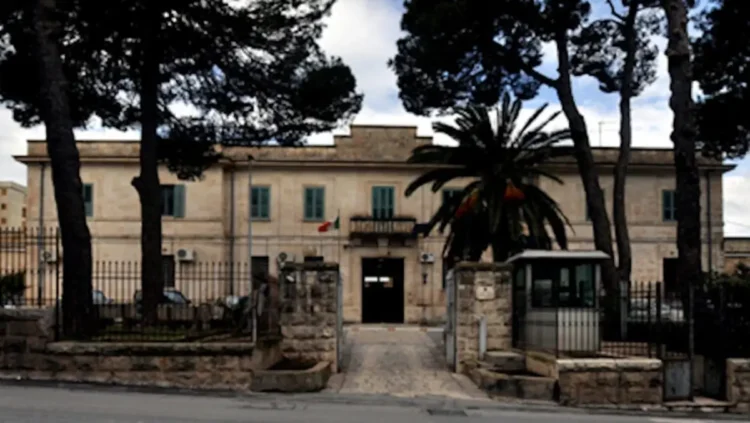 Facciata del carcere di Ragusa in pietra chiara, con cancellata, garitta all’ingresso, pini e una palma davanti