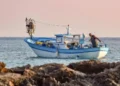 Peschereccio bianco e azzurro in mare al tramonto, con un pescatore a bordo che sistema le reti; in primo piano gli scogli
