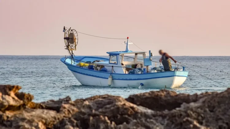 Peschereccio bianco e azzurro in mare al tramonto, con un pescatore a bordo che sistema le reti; in primo piano gli scogli