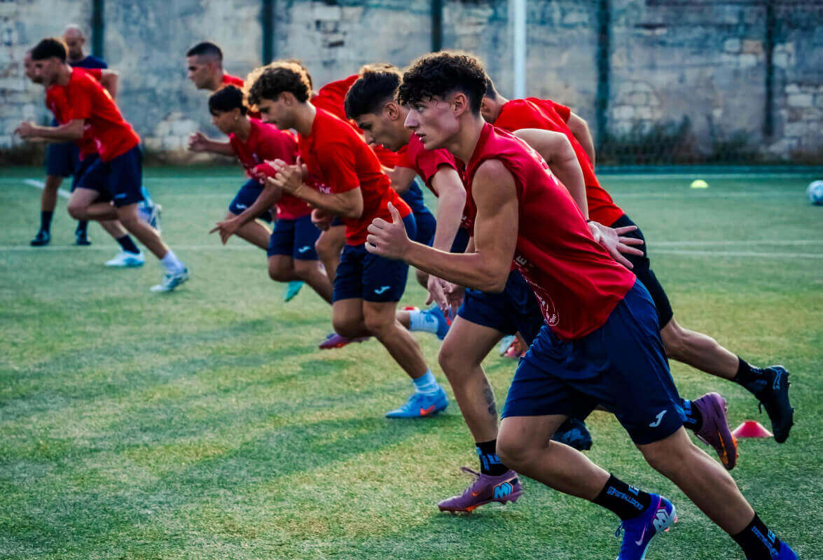 Calciatori del Frigintini con maglia rossa e pantaloncini blu scattano in fila durante un allenamento su un campo in erba sintetica