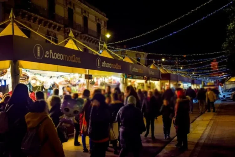 Stand di ChocoModica illuminati di sera lungo una via di Modica, con visitatori tra le bancarelle e luminarie sopra la strada