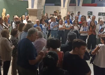 Pubblico in piedi tra archi in pietra e quadri appesi a pannelli bianchi alla mostra del Collettivo BAI a Comiso